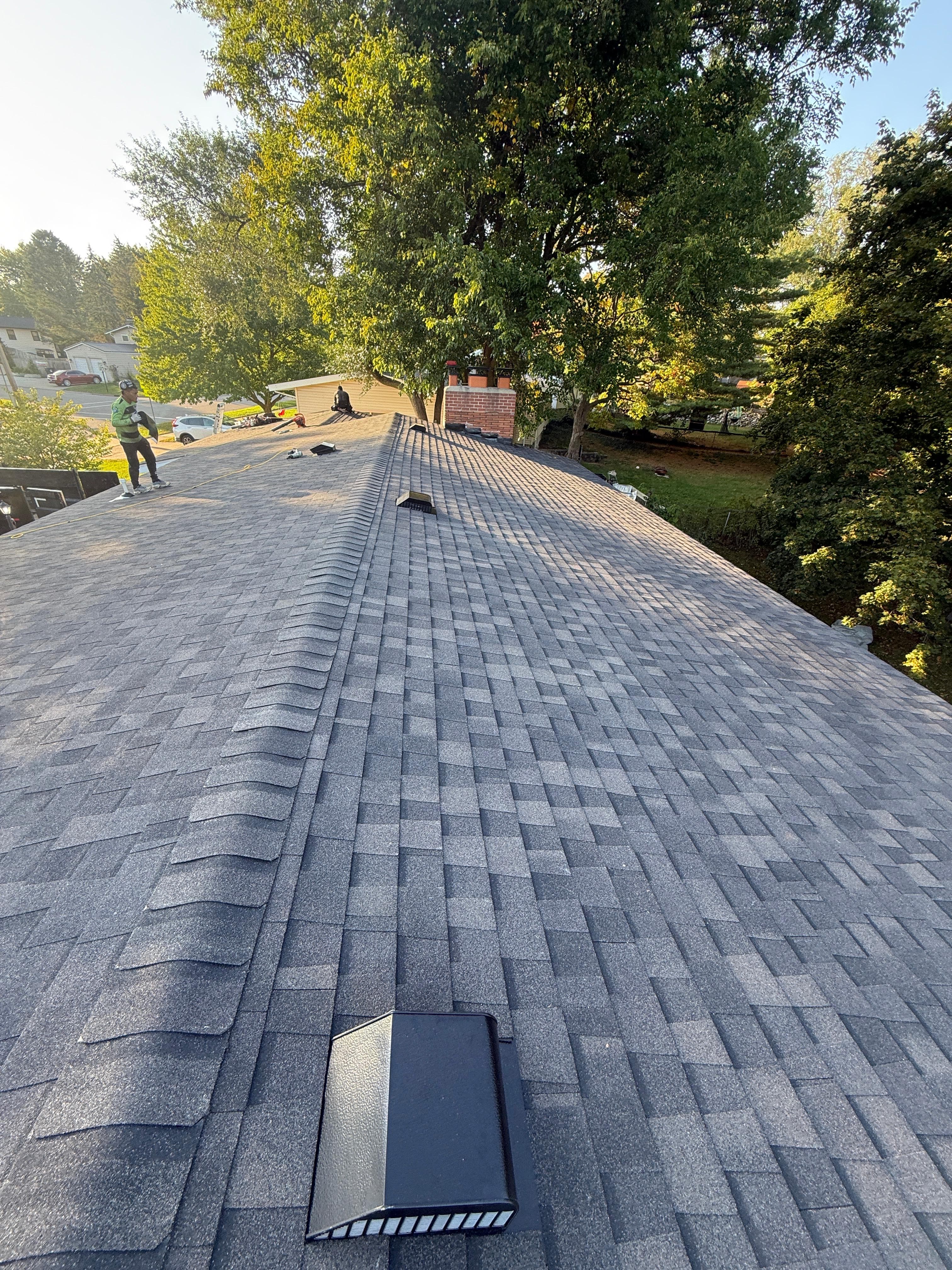 Roofers working on a large grey shingled roof peak surrounded by lush green trees.